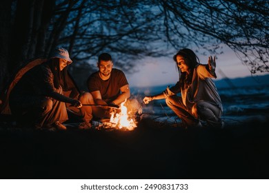 Group of happy friends enjoying togetherness while camping, preparing dinner over a campfire by a lake at night. - Powered by Shutterstock