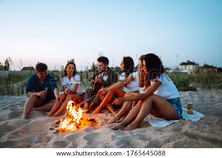 Group of happy friends frying sausages on campfire at the beach. A company of young people came together for a barbecue.
