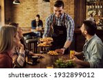 Group of happy friends having fun while waiter is serving them food in a pub. Focus is on waiter. 