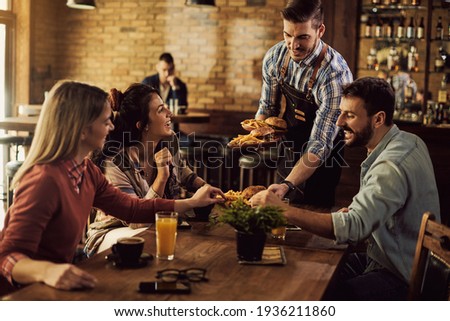 Group of happy friends having fun while waiter is serving them food in a pub. 