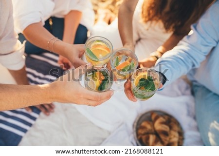 Group of happy friends having picnic, drinking lemonade and eating fruits, sitting near sea at sunset. Summer vacation.