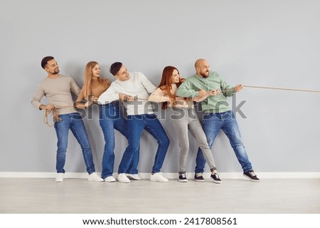 Group of happy friends playing tug of war and having fun together. Team of confident strong young adult people standing by grey color wall background, holding rope and pulling it in one direction