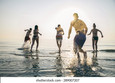 Group Of Happy Friends Running In To Water At Sunset - Silhouettes Of Active People Having Fun On The Beach On Vacation - Tourists Going To Swim On A Tropical Island