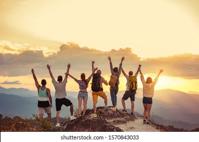 Group of happy friends stands with raised arms at sunset mountain and enjoys sunset