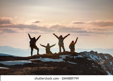 Group of happy hikers stands with raised arms in winner poses at mountain top and enjoys sunset