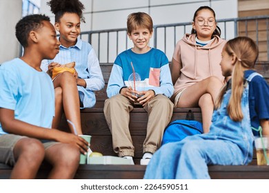 Group of happy intercultural schoolkids with snacks and soda having talk during lunch break while sitting on wooden staircase outdoors - Powered by Shutterstock