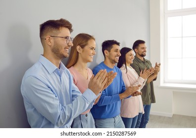 Group Of Happy Joyful Young People Applaud At Business Conference Or Team Meeting. Side View Of Smiling Modern Business Men And Women Standing In Row Near Wall And Applauding Their Leader.