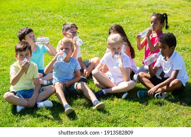 Group Of Happy Kids Drinking Water And Sitting On Grass In Park Outdoors