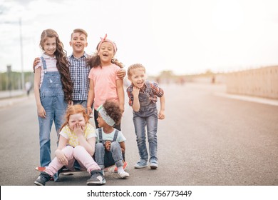 Group Of Happy Kids Outdoors. Redhead Girl And African Girlsitting On Skateboard. The Concept Of Friendship And Intercultural Communication