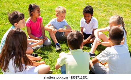 Group Of Happy Kids Resting On Grass Together And Chatting In Park At Summer Day