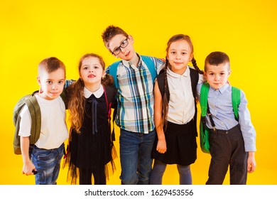 Group Of Happy Kids In Uniform With School Bags Hugging And Looking To Camera Over Yellow Background