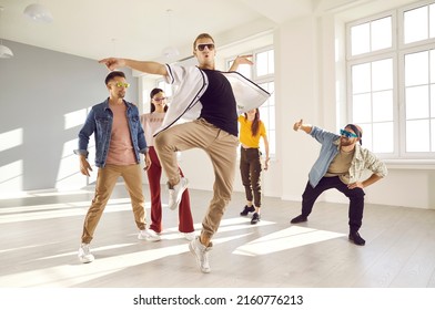 Group of happy men and women dancing in modern studio. Young male dancer wearing trendy outfit and sunglasses mixing styles and doing ballet pirouette during contemporary dance class with friends