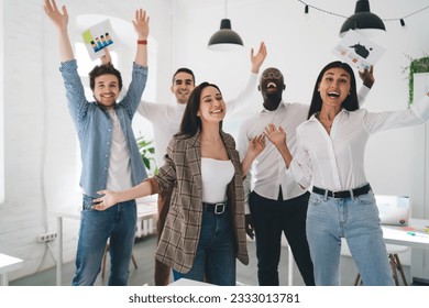 Group of happy multiethnic employees screaming dancing and raising arms celebrating victory throwing papers in air in light creative office