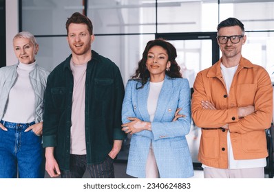 Group of happy multiracial coworkers in blurred modern office near glass wall and looking away during daytime while two colleagues standing with arms folded across chest others keeping hands in pocket - Shutterstock ID 2340624883