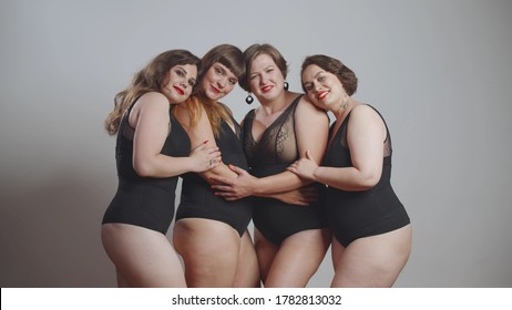 Group Of Happy Oversize Women In Black Bodysuits Standing Together Over Grey Background. Four Smiling Plus Size Models Posing At Camera Isolated. Body Positive Concept