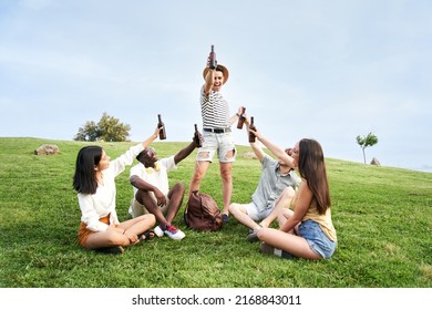 Group of happy people cheering with a beer at music festival. Five friends celebrating outdoors sitting on the grass at park.  - Powered by Shutterstock