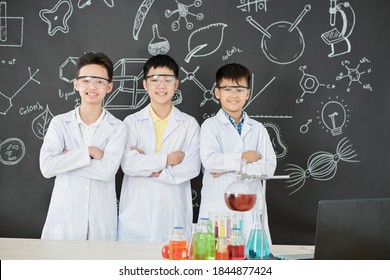 Group Of Happy Schoolchildren In Labcoats And Protective Goggles Folding Arms And Smiling At Camera