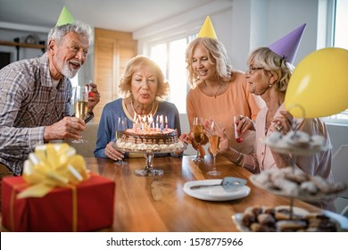 Group Of Happy Seniors Having Fun On Birthday Party While Woman Is Blowing Candles On Birthday Cake At Home. 