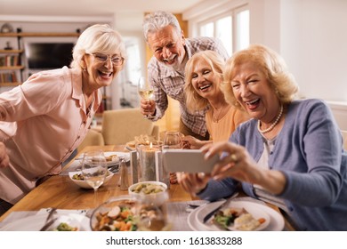 Group Of Happy Seniors Using Smart Phone And Taking Selfie At Dining Table At Home. 