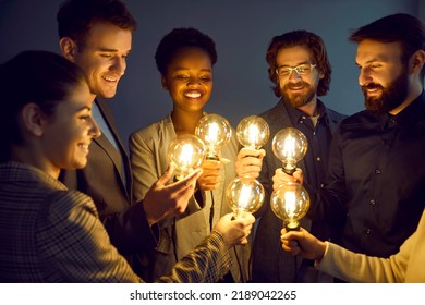 Group Of Happy Smart People Hold Bright, Shining Light Bulbs As Symbol Of Creative Ideas. Smiling Multiracial Men And Women In Business Attire Stand Together With Light Bulbs On In Dimly Lit Office.