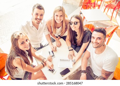 Group Of Happy And Smiling Friends Sitting In A Bar And Looking At The Camera, Many Digital Devices On The Table - Group Of Students Meet In A Cafe Outdoors