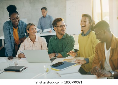 Group Of Happy Students Having Fun And Laughing While Using Laptop And Studying In The Classroom. Their Teacher Is In The Background. 