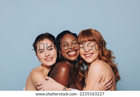 Group of happy women with different skin tones smiling and embracing each other. Three diverse women feeling comfortable in their natural skin. Body positive young women standing together in a studio.