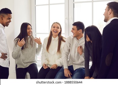 Group of happy young diverse people sitting on windowsill, telling jokes, laughing and enjoying informal communication. Coworkers, college students or friends talking about funny things during break