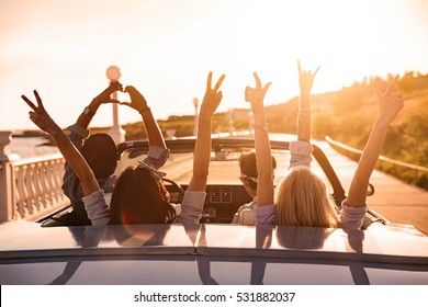 Group of happy young friends in cabriolet with raised hands driving on sunset