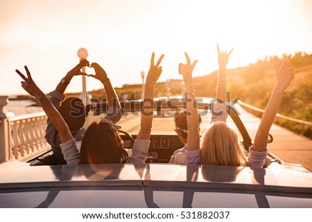 Group of happy young friends in cabriolet with raised hands driving on sunset