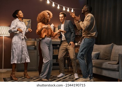 Group of happy young friends in casualewar dancing together while enjoying home party in living room illuminated by row of lamps - Powered by Shutterstock