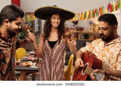 Group of happy young friends in traditional ethnic attire having fun at home party while girl in sombrero shaking maracas between guys - Powered by Shutterstock