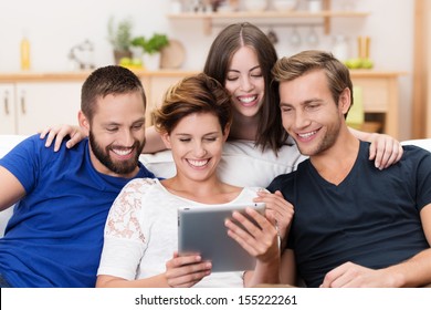 Group Of Happy Young Men And Women Sitting On A Sofa Together Sharing A Tablet Computer And Reading Information On The Screen