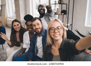 Group of happy young multiethnic coworkers in casual clothes taking selfie and looking at camera while relaxing on sofa and enjoying free time in office - Powered by Shutterstock