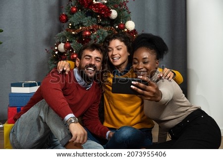 Group of happy young people best friends are sitting in front of a Christmas tree and talking on a video call via smartphone with friends who could not come to the celebration. Holidays concept.