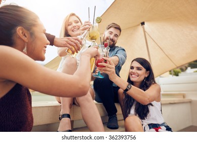 Group of happy young people cheering with cocktails and smiling while partying together on rooftop. Multiracial young friends hanging out.