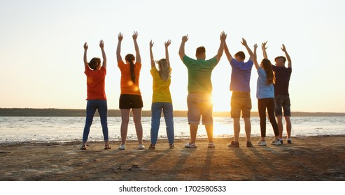 Un grupo de jóvenes felices en pantalones coloridos saltan con las manos levantadas y alegrías en la orilla del río, levantando las manos en los rayos del sol poniente.