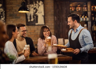 Group of happy young people drinking beer while waiter is brining food at their table in a tavern. 