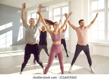 Group Of Happy Young People Enjoying A Cardio Fitness Workout Class At The Gym. Cheerful Motivated Friends Willing To Stay In Good Shape Doing Jumping Jacks With A Professional Sports Instructor