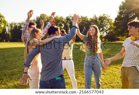 Group of happy young people have fun together while walking in park on warm summer evening. Cheerful excited multiethnic friends laugh out loud and give each other high five.