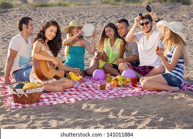 Group of happy young people having a picnic on the beach. Group of young people having an enjoyable picnic on the beach with healthy food, three of them playing music, young female is playing guitar. - Powered by Shutterstock