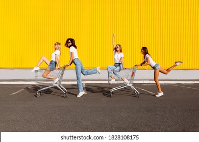 Group of happy young people having fun on shopping carts. Young beautiful women having fun and riding trolleys on a sunny summer day against the background of the yellow wall of the shopping mall