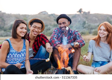 Group Of Happy Young People Sitting And Roasting Marshmallows On Campfire Outdoors