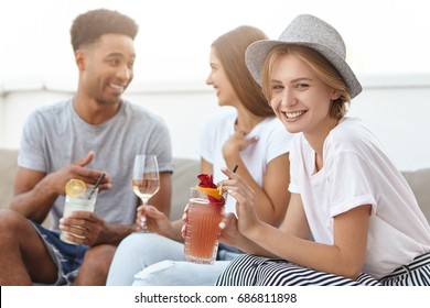 Group Of Happy Young People Toasting To Love And Prosperity, Having Alcohol Drinks At Rooftop Party. Three Cheerful Friends Enjoying Nice Time Together, Having Fun At Terrace Cafe, Drinking Cocktails