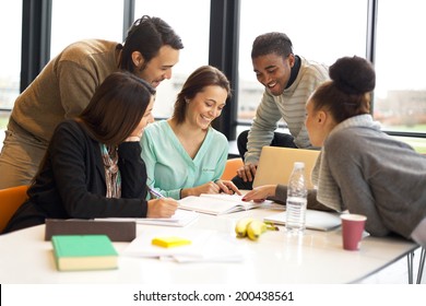 Group Of Happy Young Students In Cooperation With Their School Assignment. Multiethnic Young People Sitting At Table Reading Reference Books For Study Notes.