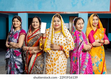 Group of happy young traditional indian women housewives wearing colorful sari showing smart phone with blank display screen to put advertisement, Rural india. women empowerment.