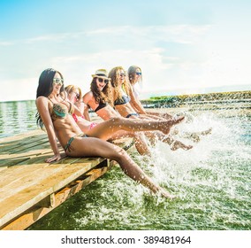 Group Of Happy Young Woman Feet Splash Water In Sea And Spraying At The Beach On Beautiful Summer Sunset Light. Five Sexy Girls Playing On Wooden Pontoon Against Blue Sky Background Enjoy Holiday