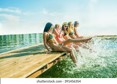 Group Of Happy Young Woman Feet Splash Water In Sea And Spraying At The Beach On Beautiful Summer Sunset Light. Five Sexy Girls Playing On Wooden Pontoon Against Blue Sky Background. Enjoy Holiday