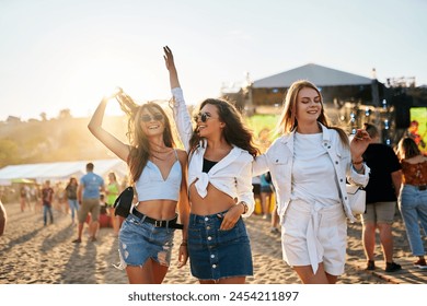 Group of happy young women dance at a beach festival, sunset lights their carefree moves. Friends in summer attire enjoy music, sandy shores, sea breeze. Casual outdoor event, youth culture, freedom.