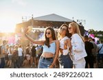 Group of happy young women friends take selfie at sunny beach music fest. Girls in trendy summer outfits enjoy live concert, party atmosphere. Youth culture, friendship, outdoor entertainment moment.